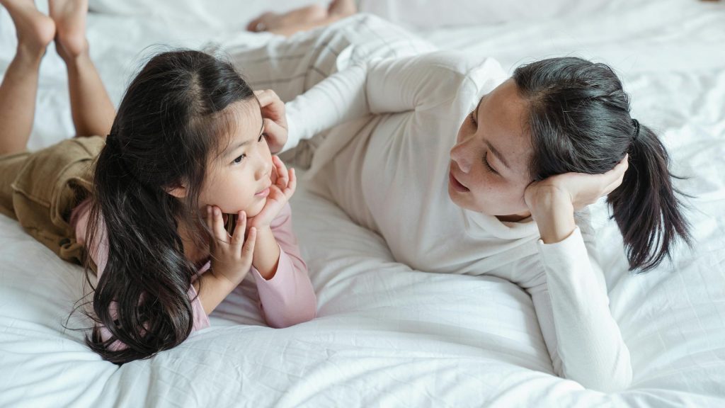 A mother and daughter bonding indoors, lying on a bed and enjoying quality time together.